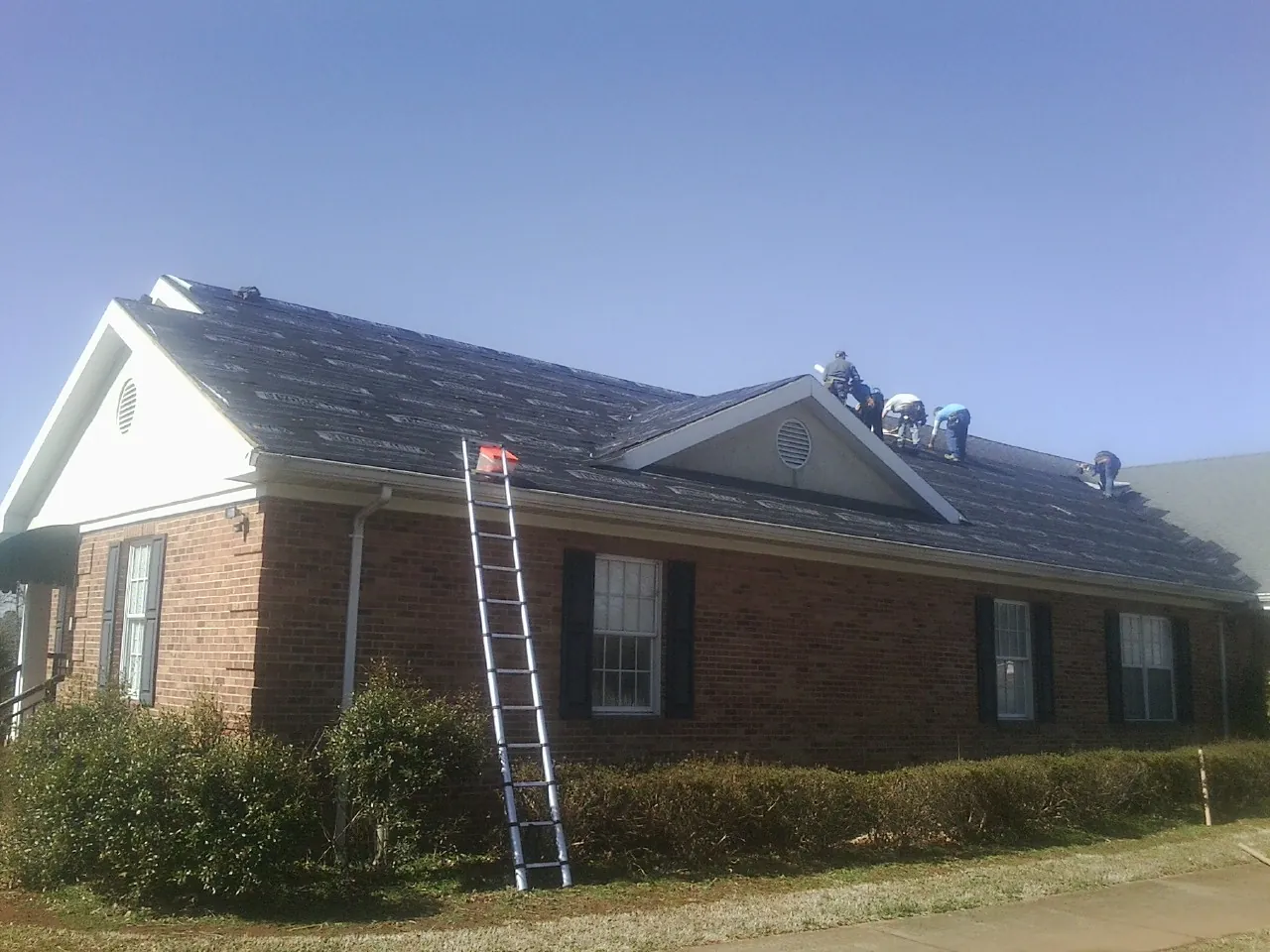 Crew tearing shingles off a brick ranch roof with a ladder set — Emergency Roof Repair work by our crew in Driftwood
