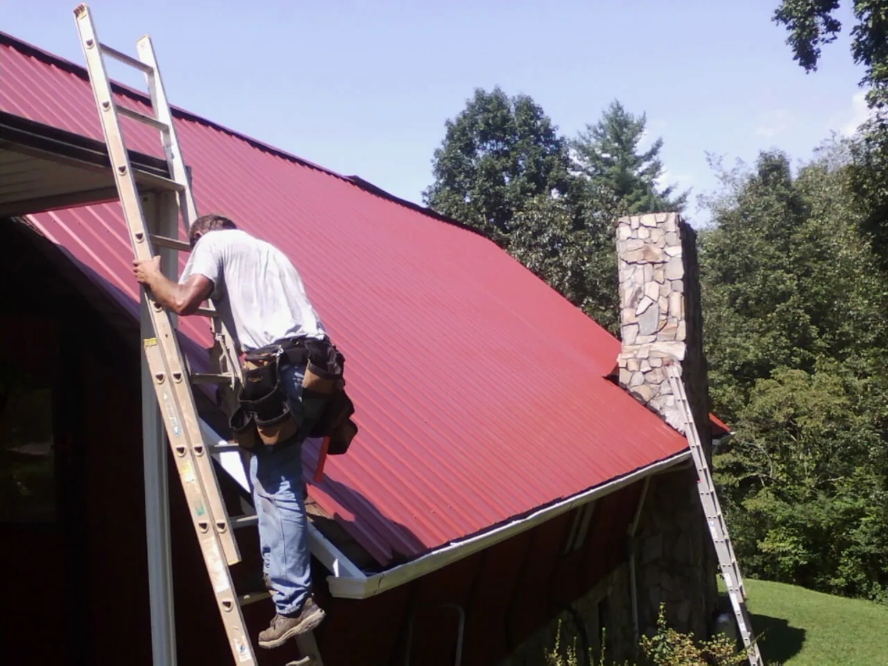 Roofer on a ladder beside a red metal roof and stone chimney — Soffit & Fascia Repair by Roofing Brothers in Driftwood